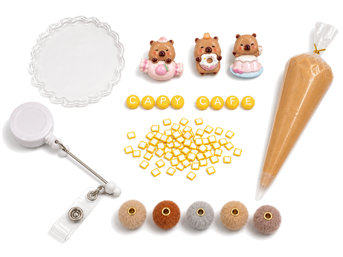 CAPY CAFE DIY badge reel kit with three capybara charms, CAPY CAFE letters, toast pieces, neutral pom-pom beads, reel and caramel cream glue on a pure white background